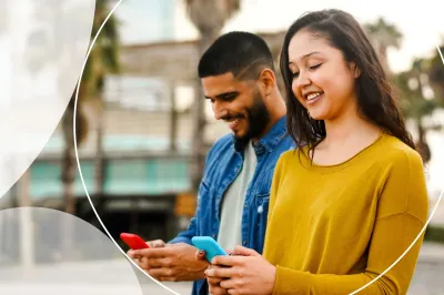 Man and woman smiling and looking at phones with background of buildings and palm trees.