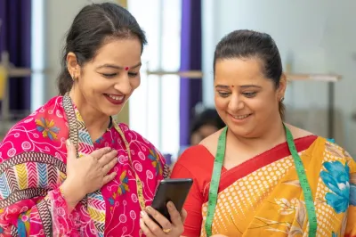 Two women in Indian dress with measuring tapes around their shoulders smiling at a phone.