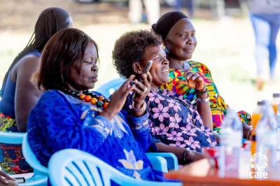 A woman in a vibrant blue dress uses a smartphone during a Village Savings and Loan Association (VSLA) meeting, as fellow members look on with interest, at an outdoor community gathering in Africa
