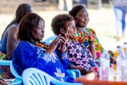 A woman in a vibrant blue dress uses a smartphone during a Village Savings and Loan Association (VSLA) meeting, as fellow members look on with interest, at an outdoor community gathering in Africa