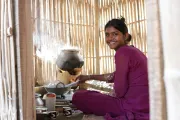 Woman in Nepal sitting at a stove cooking within a bamboo enclosure, smiling at the camera.