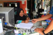 Indonesian woman in headscarf behind display counter with phone in hand while customer hands cash.