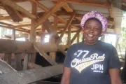 Smiling woman with hair cover standing in front of wooden structure.